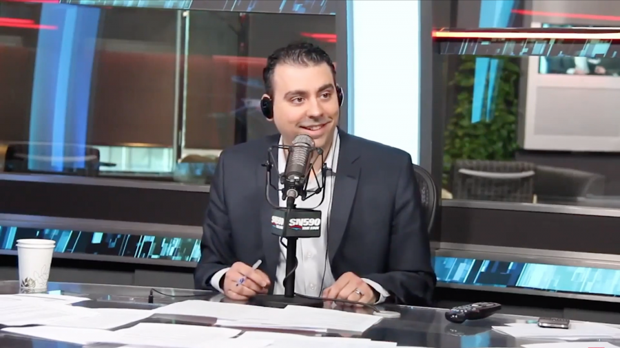 Sid Seixeiro sits at a news desk in a studio in front of a  microphone with a SN590 flag
