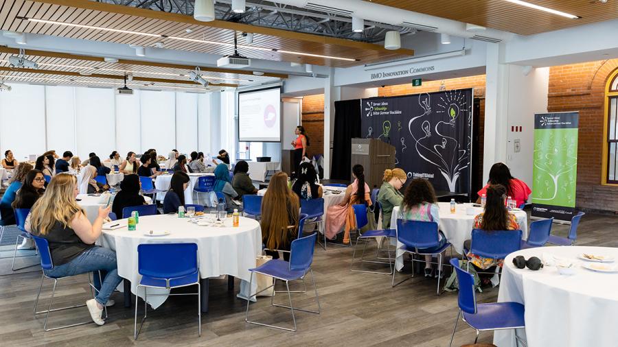 A person stands at the front of the room and is speaking while people sitting at tables listen.