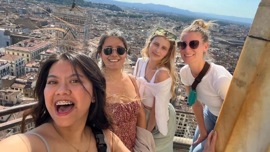 Four students smile and take a selfie with an Italian city in the background.