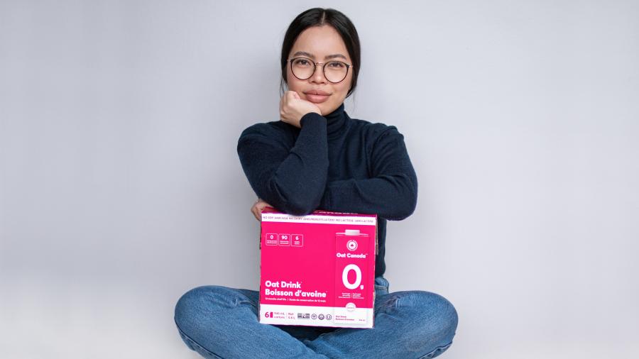 Eloise Ambursley sits cross-legged with her arms crossed atop a pink box of Oat Canada oat milk