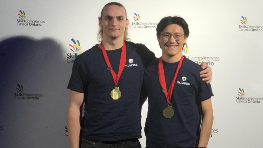 Two smiling Humber College students wear gold medals around their necks.