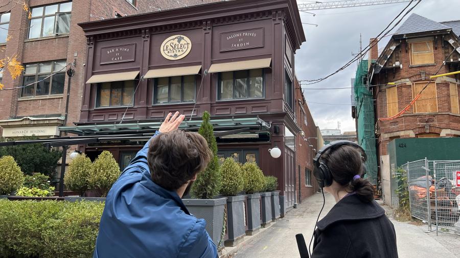 Two people holding recording equipment look towards a building with a sign on it reading Le Sélect Bistro.