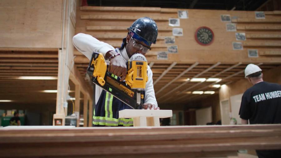 A person wearing a hardhat uses a tool to nail pieces of wood together.