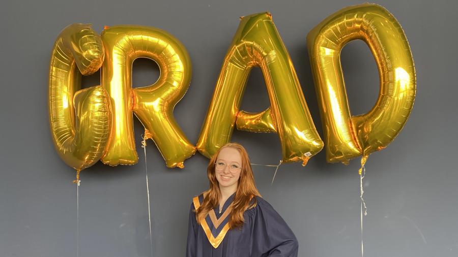 Emma Kilgannon wears a convocation gown and stands beneath balloons that spell out the word grad.