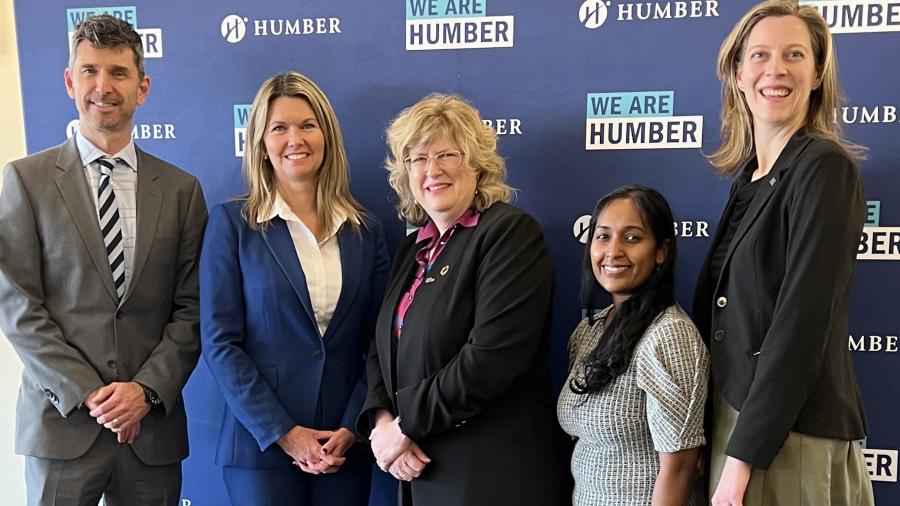 Five smiling people stand in front of a blue backdrop with the Humber College logo on it.