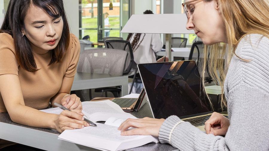 Two people are looking at a book while sitting together at a desk.