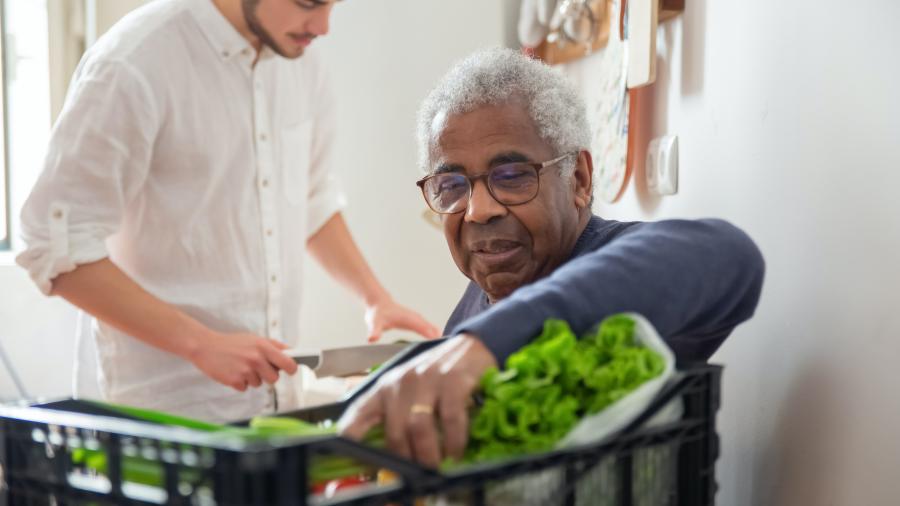 An older adult with grey hair picks up vegetables while a younger person in the background uses a knife to chop them.
