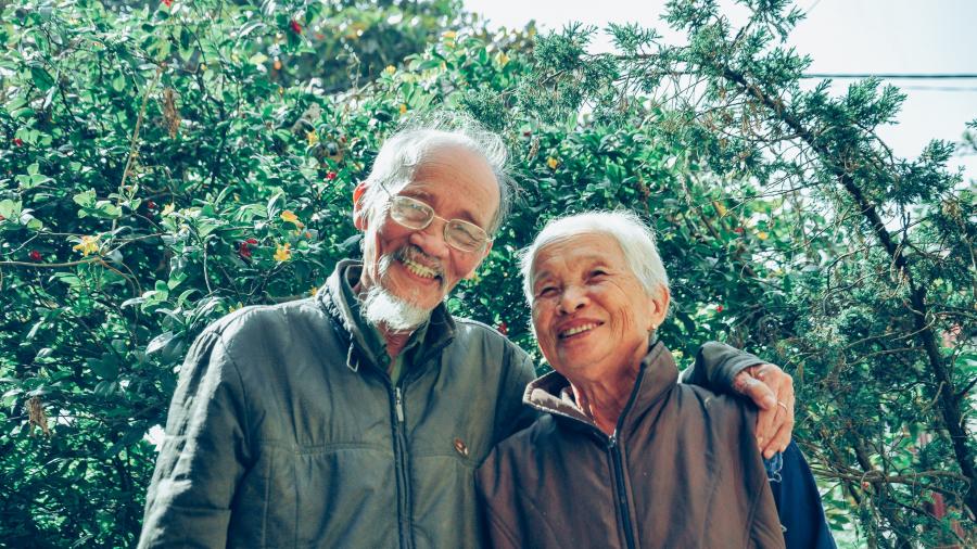 A photo of two smiling older adults standing in front of trees. One person has their arm around the other.