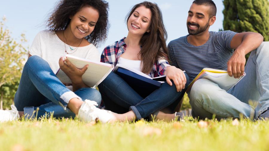 Three students sit on the grass while looking at a book