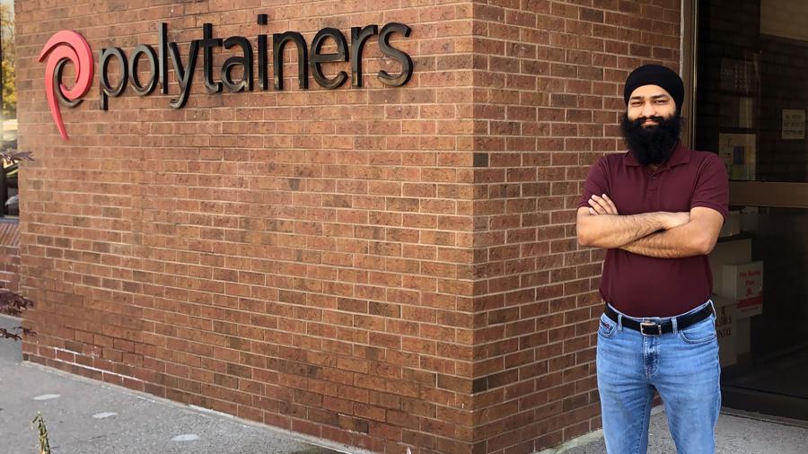 Taranjeet stands with his arms crossed beside a brick wall the the Polytainers name and logo on it.