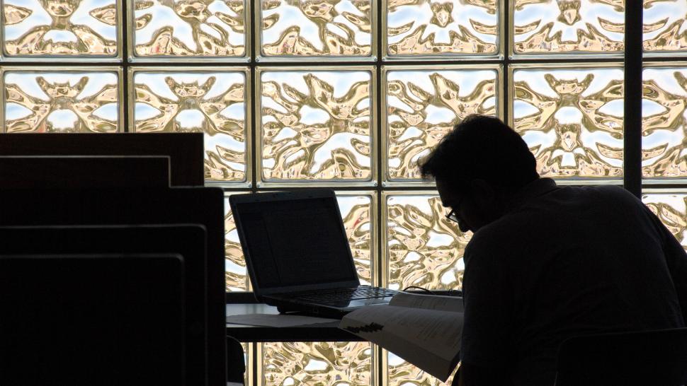 A person sits at a table working on a laptop. They are hunched and outlined in shadow in front of a glass wall.