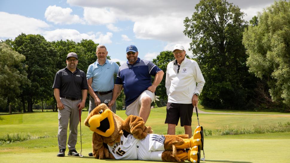 Four men stand in a row wearing golf clothes and holding golf clubs. The Humber Hawk mascot lies on the ground in front of them.