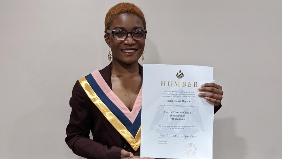 Claire Doyle holds her diploma in front of her wearing a plum-coloured suit and a convocation collar