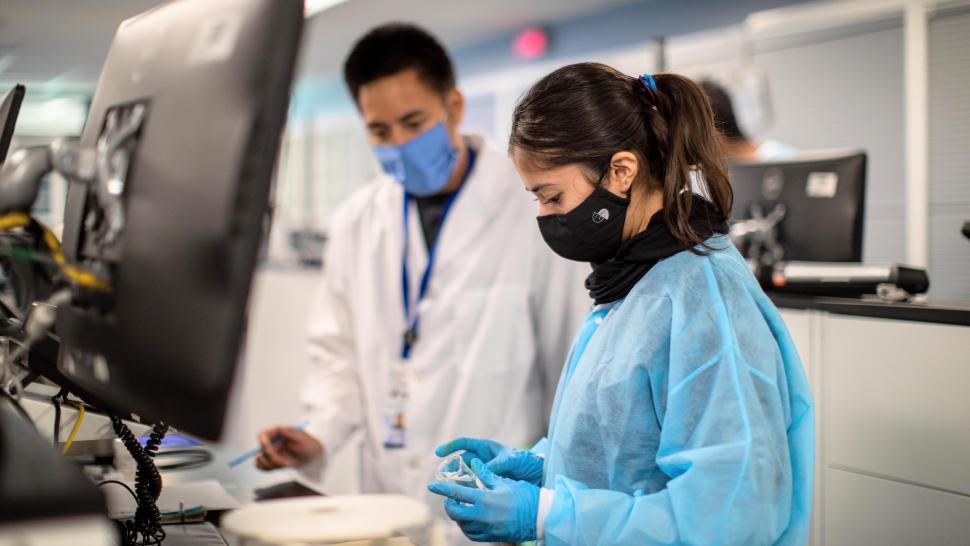 Two people in a pharmacy lab, wearing masks, gowns and gloves. 