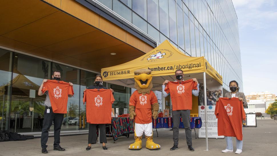 Five people stand outside of a large building, holding up orange shirts.