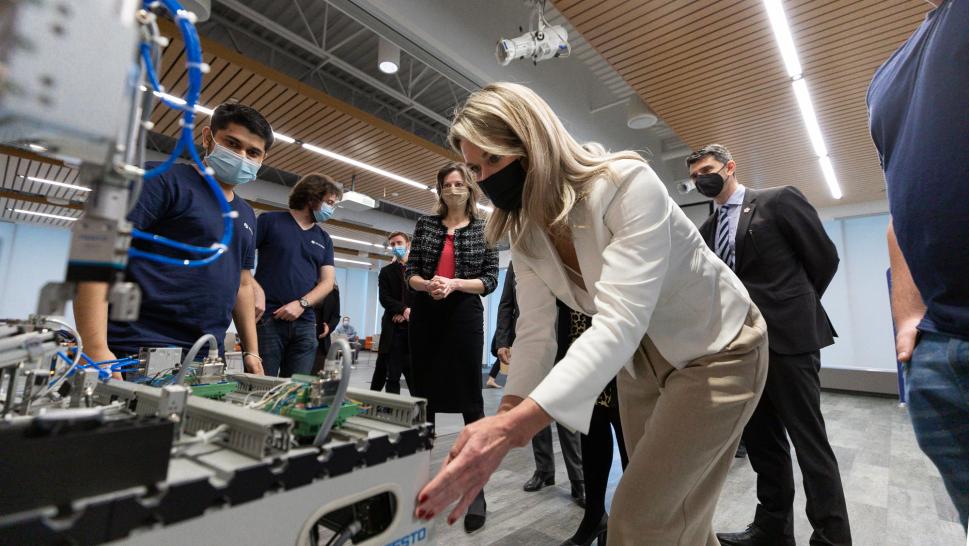 Masked MPP Jill Dunlop bends over a large, FESTO-branded machine with students and officials in the background, all masked