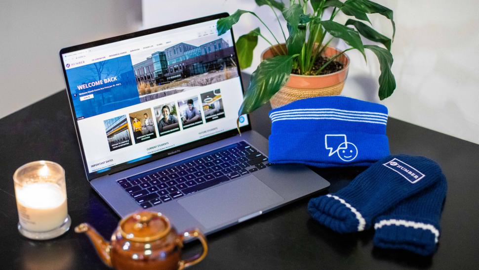 A blue hat with the BellLetsTalk logo is perched on the side of laptop keyboard. The screen shows Humber's homepage.