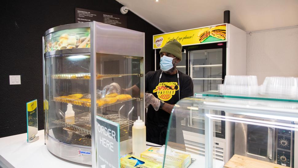 A person removes a Jamaican patty from a warming machine while wearing a t-shirt that reads Jamaican Spiked Patties.