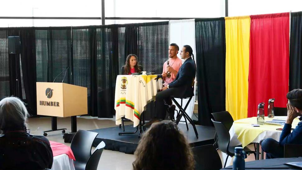 Three people sit around a table as one speaks into a microphone as a crowd listens.