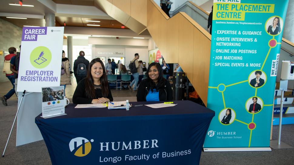 Two people sit at a desk that has a sign on it that reads Humber Longo Faculty of Business.
