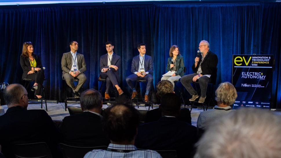 Several people sit on a stage while one person speaks into a microphone during a panel discussion.