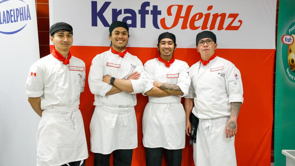 Four smiling people wearing chef’s outfits stand in front of a sign that reads Kraft Heinz.