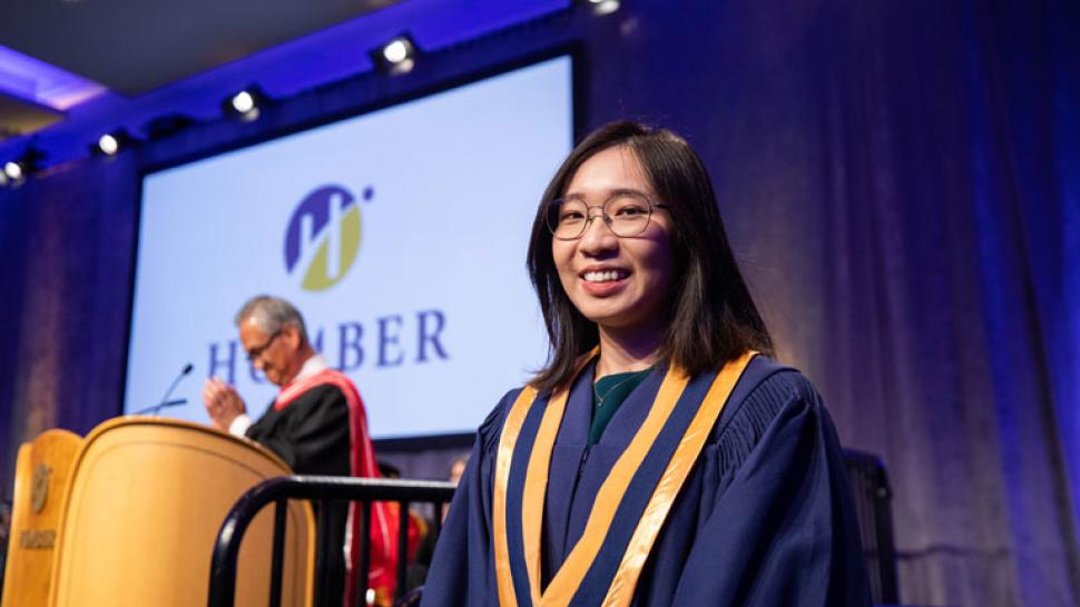 A person wearing a convocation gown smiles. In the background is another person at a podium.