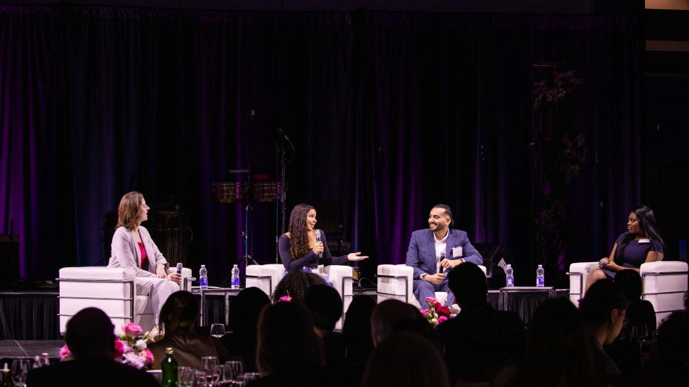 Four people holding mircophones engage in a discussion while sitting on a stage.