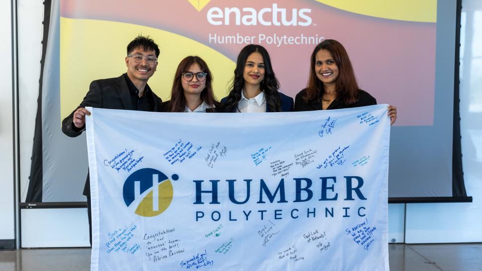 Humber Enactus Team - Reshade  Humber Enactus team of four people hold a Humber Polytechnic flag signed by supporters