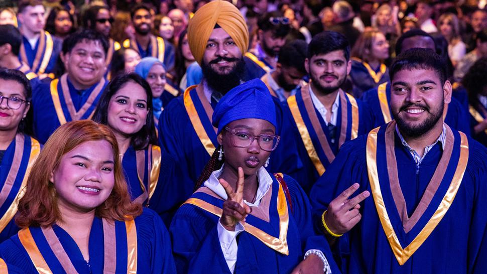 A group of graduating students wearing convocation gowns are smiling and waving.