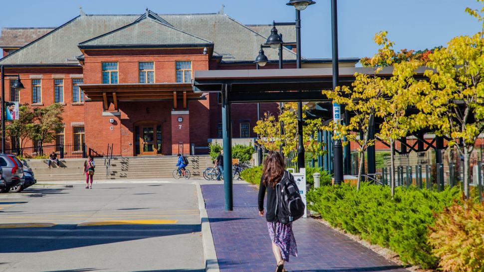 Humber College students walk on a brick sidewalk lined with trees towards one of the cottages at Lakeshore Campus on a sunny day