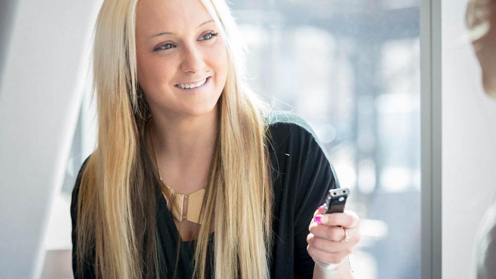 A reporter  holds a recorder up in her hand. She has long blonde hair and is smiling. She is backlit by a wall of windows