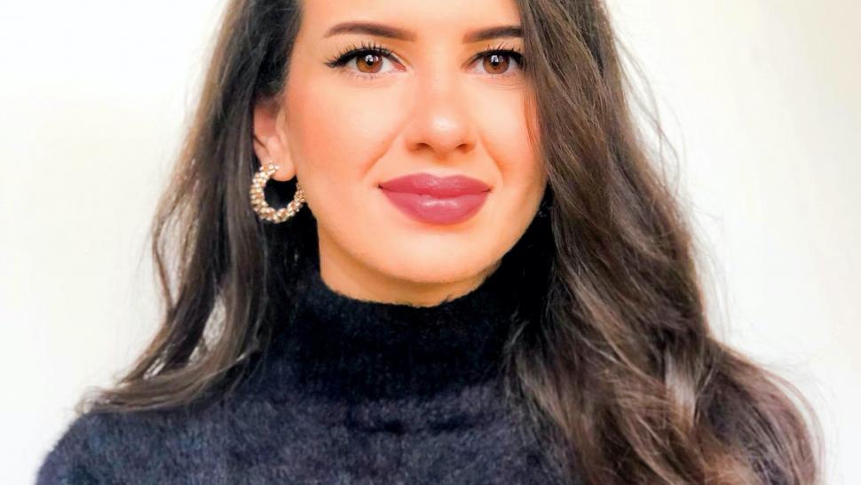 Jodi Al Hayek smiles slightly,posed in front of a white background wearing a black turtleneck sweater. She has long dark hair