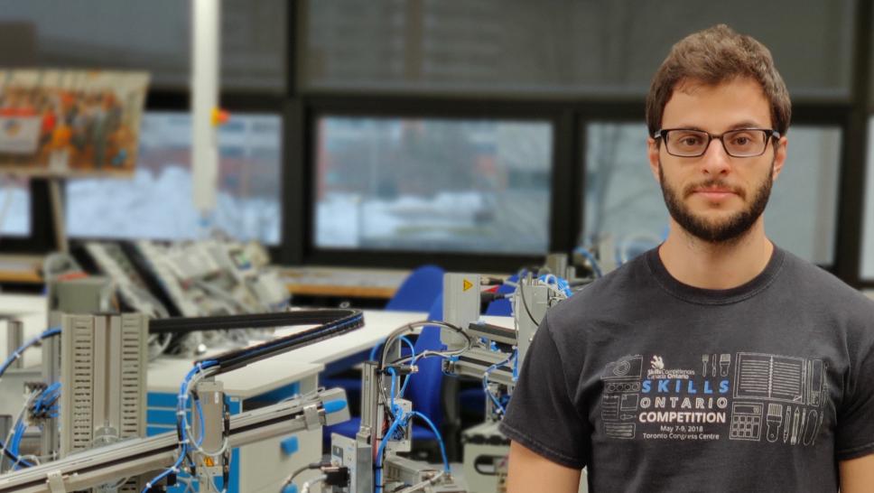 Mauricio Toigo stands in a lab at the Barrett CTI with machinery behind him. He is wearing a t-shirt, jeans and glasses