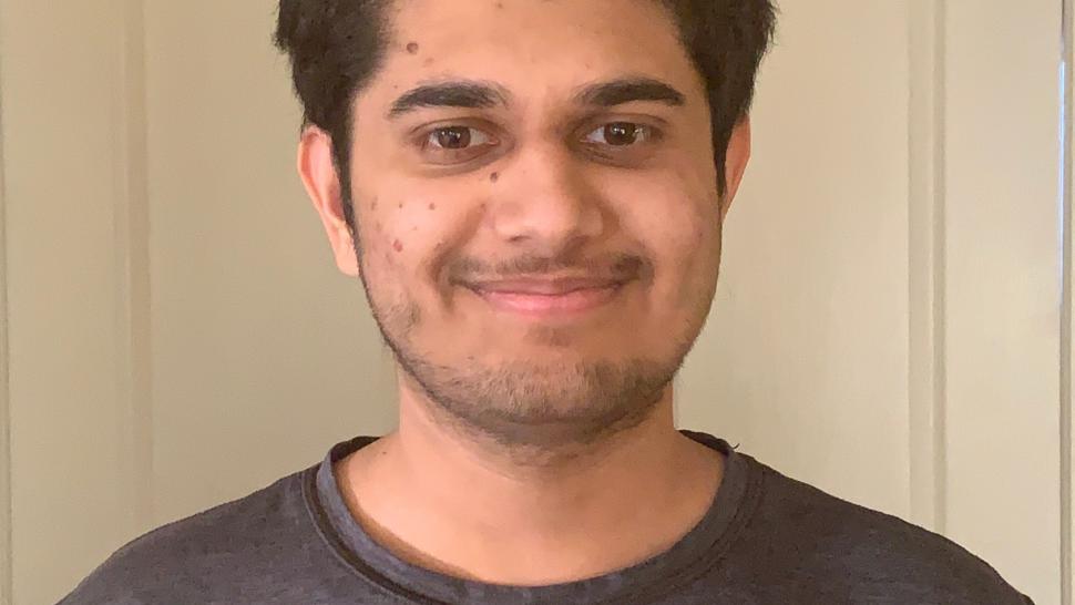 Daneep Lahl smiles at the camera with his mouth closed, standing in front of a beige wall