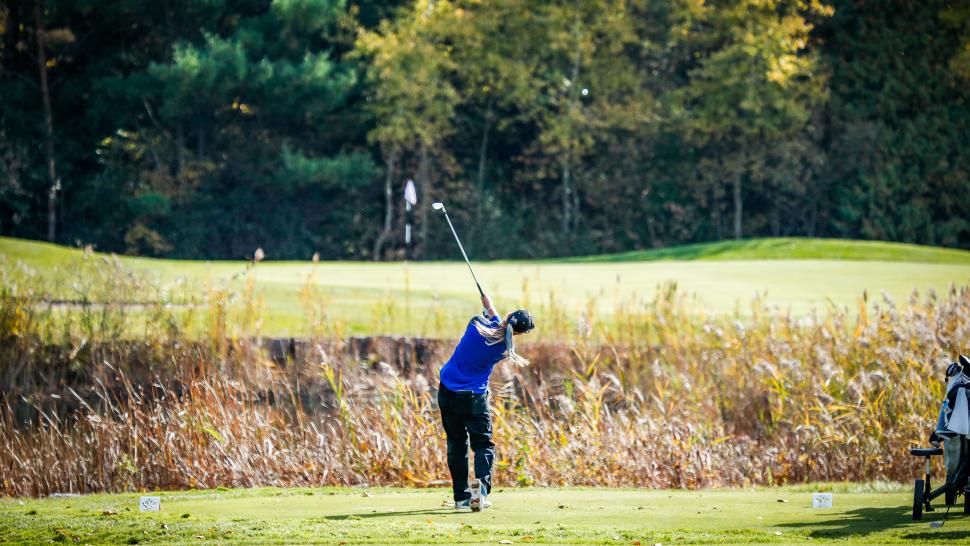 A golfer in mid-swing watches their golf ball head towards the green, which can be seen in the distance.