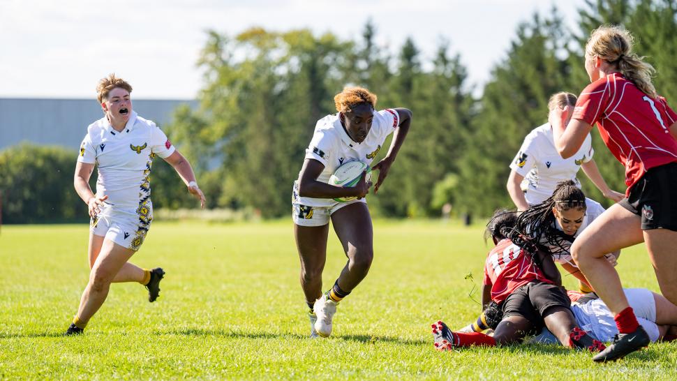 A person wearing a Humber College rugby jersey carries a ball while a teammates runs beside them. 