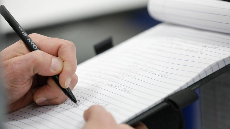 A hand is holding a black pen, writing on a small notebook, held by the other hand.