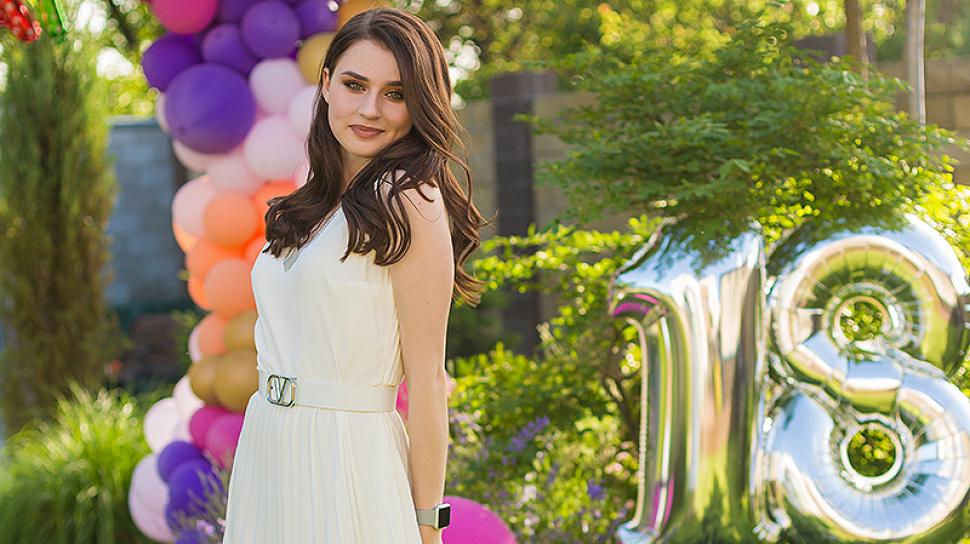 Renata Borsuk smiles softly at the camera, looking over her shoulder with balloons in the background wearing a white dress