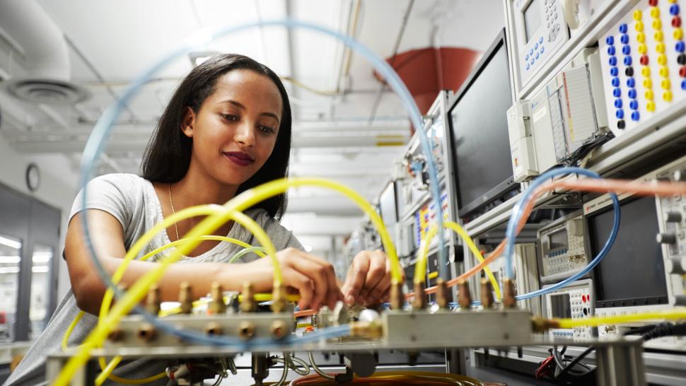 A young woman in the background connects yellow wires to a circuit board in the foreground.