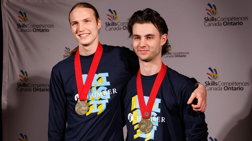 Two people pose for a photo while wearing medals and Humber shirts.