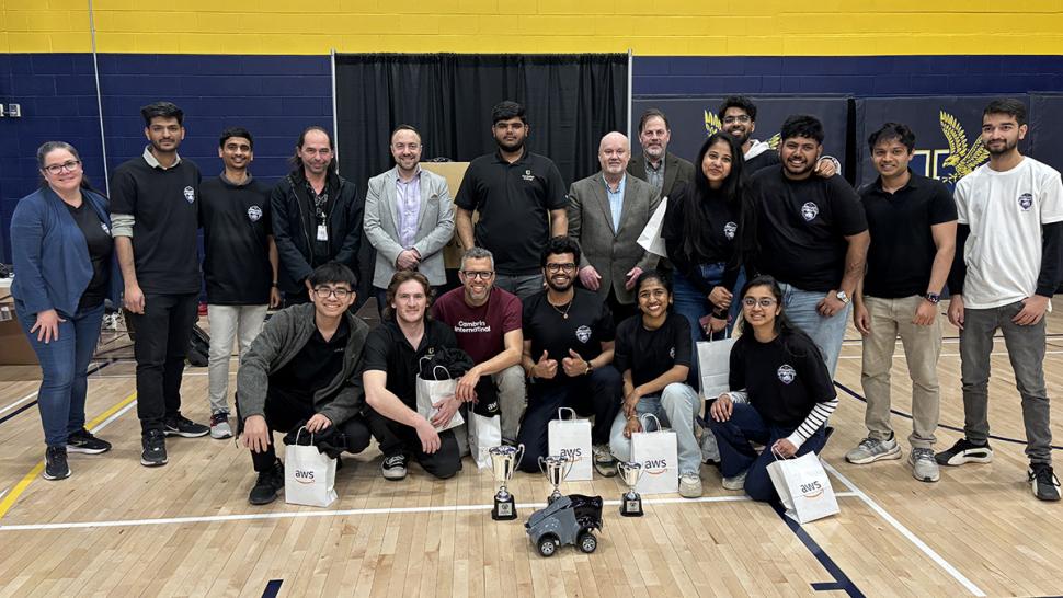 Humber Polytechnic hosted its annual AWS DeepRacer competition, which is a full autonomous racing league with a 1/18th scale race car driven by a machine learning algorithm. A large group of smiling people pose for a photo. Some have gift bags and tophies with them.
