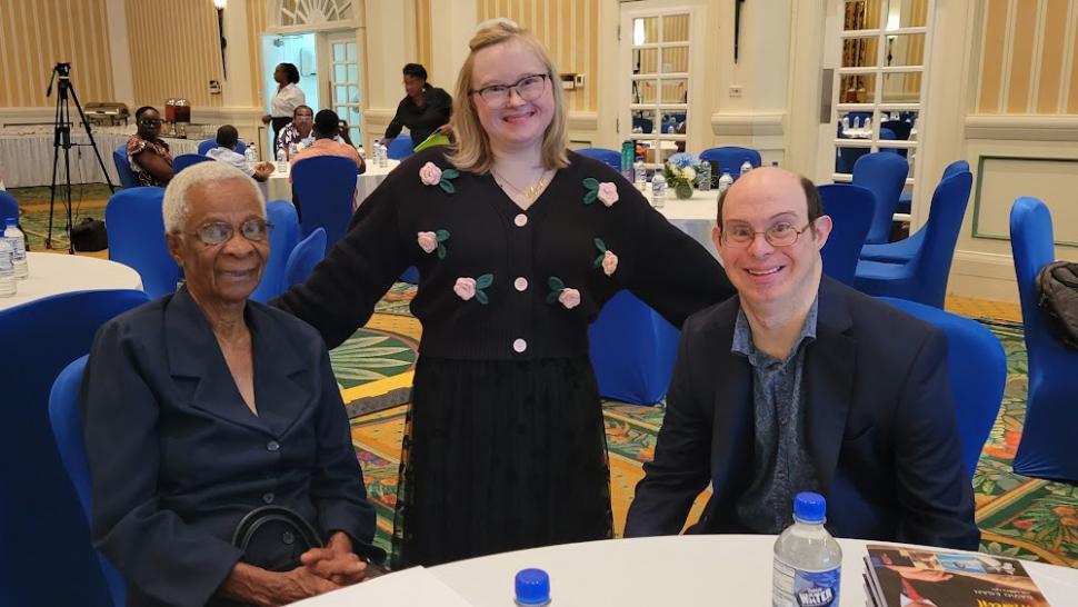 Angel Blainey is a gradute of Humber Polytechnic’s Community Integration through Co-operative Education program. A person stands between two other people who are sitting at a table.