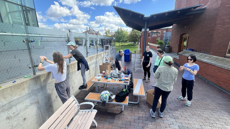 People are standing outside near a table with materials on it. Some people are working on a piece of glass. 