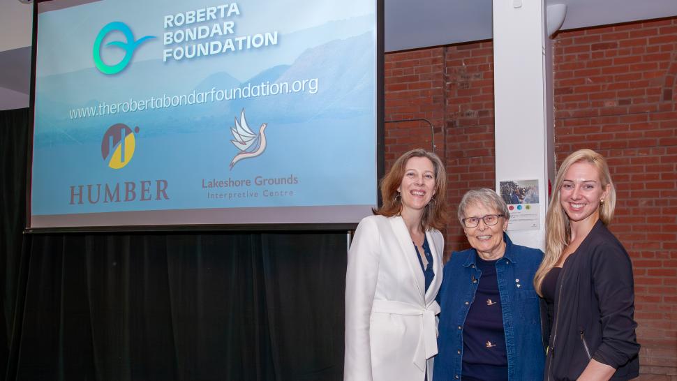Three smiling people stand beneath a screen that reads Roberta Bondar Foundation.