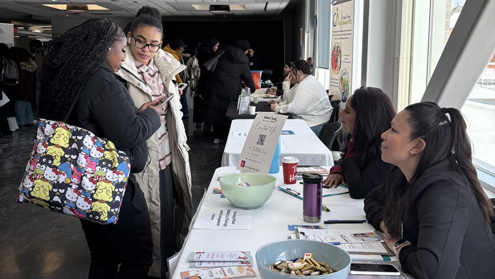 Two people stand in front of a table that has two other people sitting at it. The table has literature and other items on it.