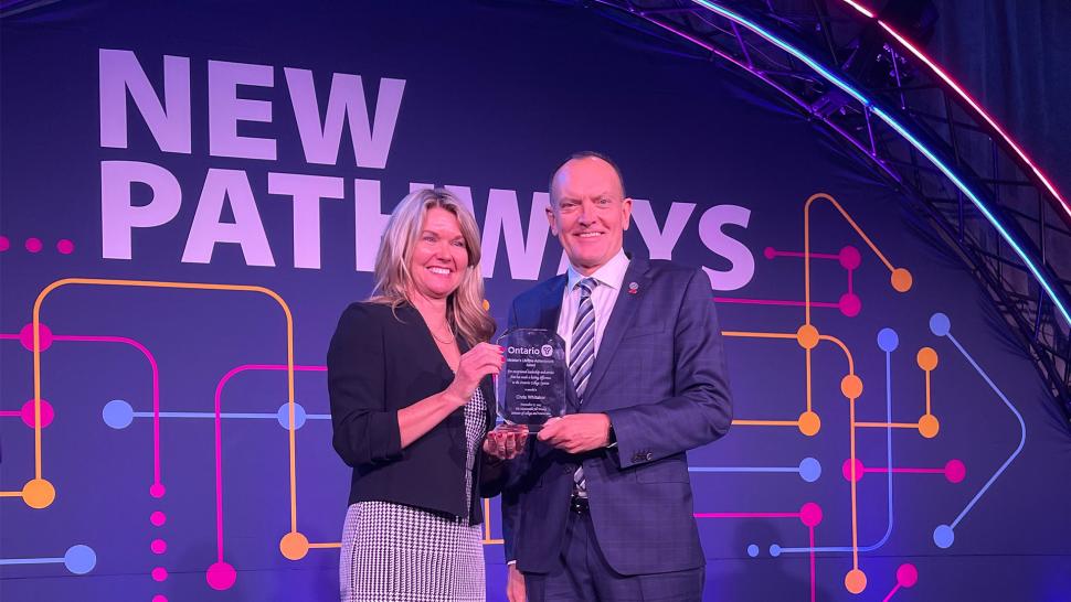 Two smiling people stand on a stage holding an award. A screen in the background reads New Pathways.