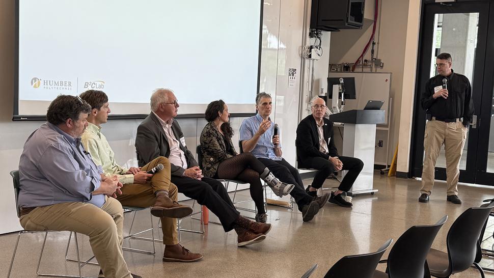 Several people sit in chairs in front of a screen and one is speaking into a microphone.