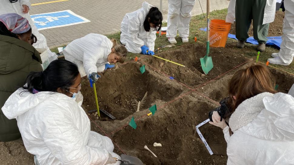 Students wearing white jumpsuits document a simulated gravesite where fake bones can be seen in the dirt.