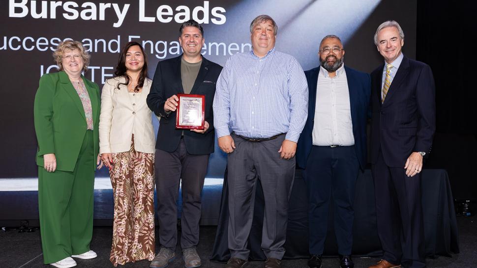 Several people stand together on a stage. One is holding an award.
